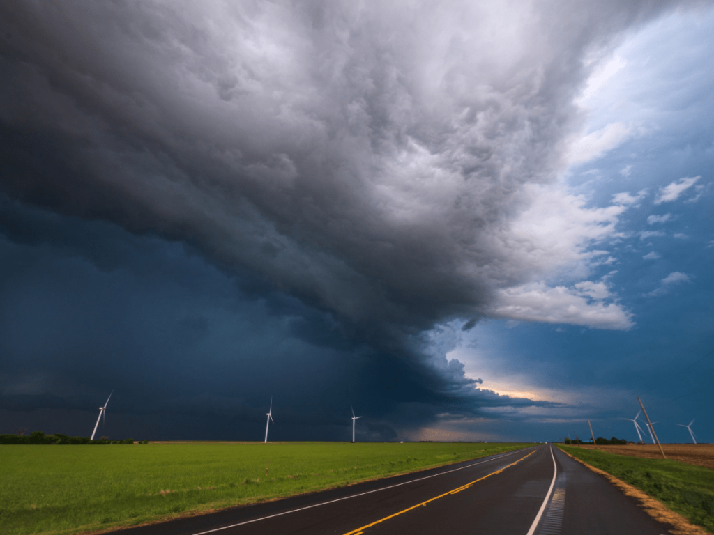 A road near a green field with dark clouds signaling bad weather above.