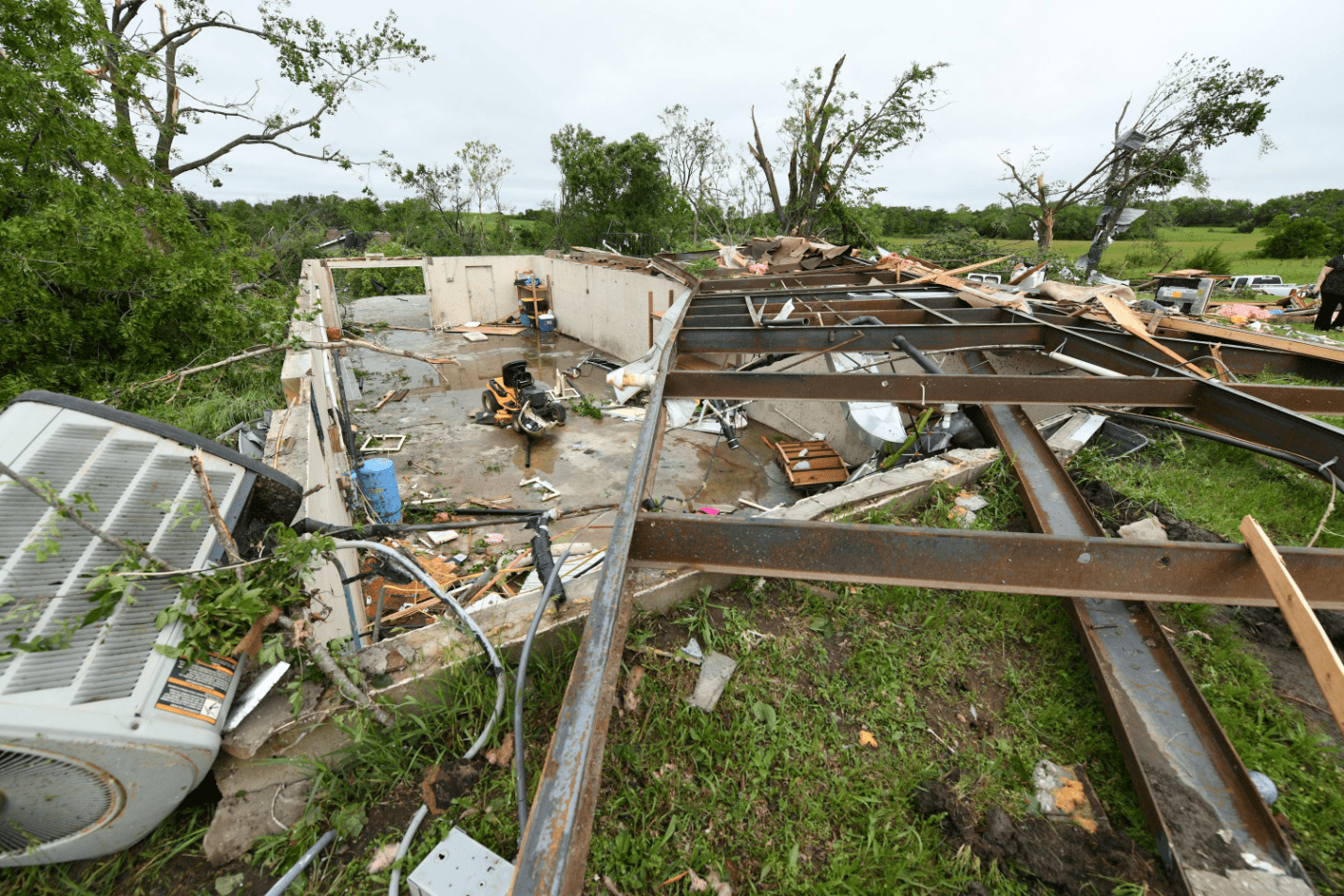 A broken, partially collapsed house with debris lying around it sits abandoned in a green field under a cloudy sky