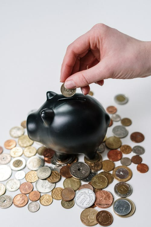 A hand putting a coin inside a black piggy bank surrounded by coins