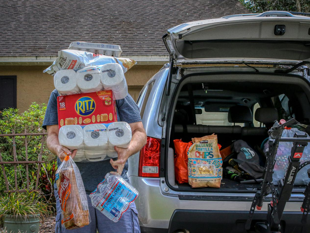 A man standing outside the open trunk of a car holding supplies, such as toilet paper