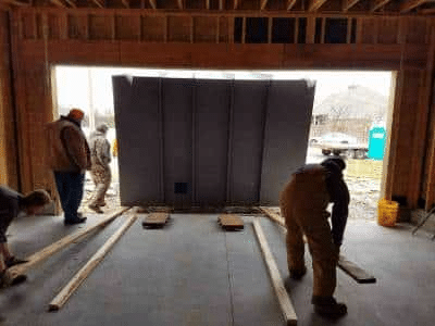 A picture shows two workers installing a gray, modular storm shelter inside a framed building.