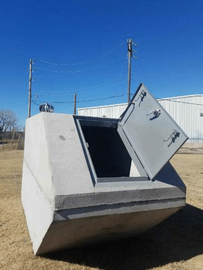 An image shows a pre-fabricated concrete storm shelter with its metal door open in an outdoor setting
