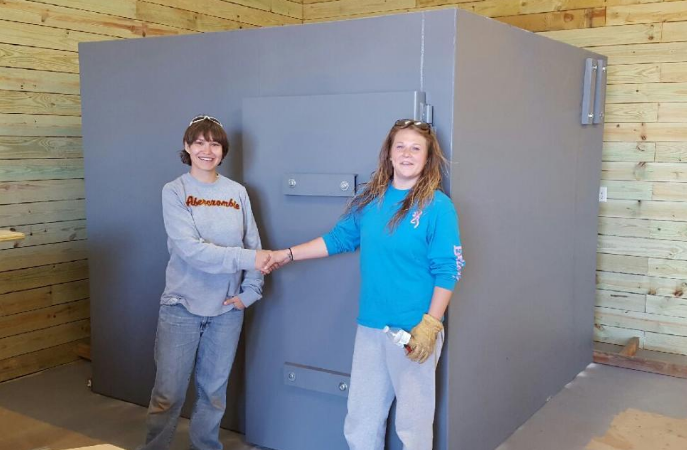 Two women shake hands in front of a grey safe room that’s been installed.